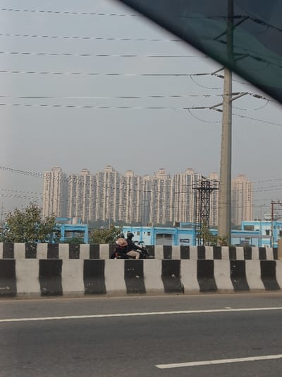 This image captures an urban landscape in Ghaziabad, India, featuring a blend of infrastructure, residential buildings, and daily activity under a hazy sky. The photograph appears to be taken from a moving vehicle, with a dark section of a car window or roof visible in the upper right corner.
In the immediate foreground, a wide asphalt road is visible. A prominent concrete barrier with alternating black and white stripes runs across the midground, separating the main road from a lower-level path or service road. On this path, two individuals are seen riding a motorcycle. The driver wears a dark helmet and jacket, and the passenger, dressed in lighter attire, has a brown backpack or parcel. Sparse green foliage and bushes are visible behind the barrier to the left.
Behind the barrier, a series of low-rise buildings with blue exterior walls and white roofs are present. Text, possibly reading "LAPP OMN," can be faintly discerned on one of these blue structures. To the right, a tall, grey utility pole stands, supporting multiple power lines that extend horizontally across the frame.
Dominating the background is a long row of numerous tall, light-colored residential apartment buildings. These multi-story structures exhibit similar architectural styles with many windows and balconies, forming a significant urban development against the horizon.
The sky is a uniform, pale grey, suggesting an overcast day or potentially hazy/smoggy atmospheric conditions, common in the region. The scene depicts a typical daytime urban environment with ongoing movement and extensive building development.