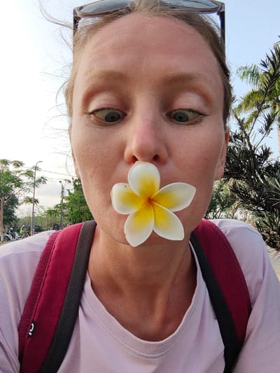 A close-up portrait shot in Thuận Hóa Ward, Vietnam, features an adult female subject from the chest upwards. She has fair skin and light-colored hair, with dark sunglasses resting on top of her head. Her expression is characterized by wide-open eyes looking downwards, and a white Plumeria flower with a yellow center is positioned between her nose and upper lip, covering her mouth. She wears a light purple crew-neck t-shirt and a maroon backpack with a dark grey padded strap, visible over her left shoulder. The outdoor background on the left includes deciduous trees, a street light pole, and several distant figures on motorbikes on what appears to be a road under a bright, overcast sky. On the right, a large palm tree and dense dark green foliage are visible.