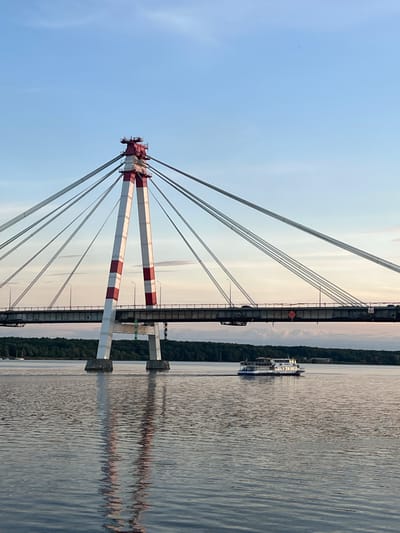 The image shows a cable-stayed bridge spanning a body of water, likely a river or lake, with a boat in the foreground. The bridge features two large, white and red striped pylons supporting numerous cables that hold up the bridge deck. Vehicles are visible on the bridge, indicating it is a functional roadway. The water reflects the sky and the bridge structure. In the distance, a treeline forms the background, with a small settlement or buildings visible on the far bank. A white passenger boat with multiple decks is moving across the water. The sky is a clear blue with some pale clouds, suggesting either early morning or late afternoon. This scene is characteristic of the Rybinsk Reservoir or the Sheksna River within the city of Cherepovets, Russia, specifically near the Cherepovets Bridge (also known as the Northern Bridge). There are no visible people.