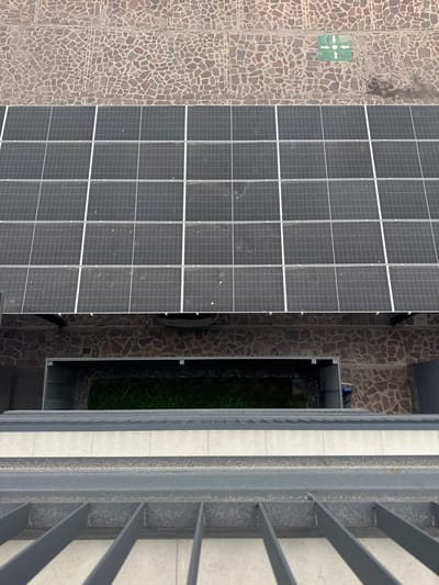 The image captures a high-angle, downward view of an urban environment in León, Mexico. In the immediate foreground, a gray metal railing with horizontal bars is partially visible. Directly below the railing, a lower-level courtyard or recessed area contains a rectangular patch of green grass, enclosed by dark-colored walls and irregular brown stone paving. A partially visible blue object, possibly a container, is situated on the right side of this lower level. Above the courtyard, an array of at least twenty dark gray photovoltaic solar panels, delineated by white grid lines and exhibiting surface dust, is installed on a rooftop or elevated platform. In the uppermost background, a ground-level area paved with irregular brown cobblestones displays a green square symbol featuring white arrows pointing outwards from a central point. No individuals or active interactions are present in the scene.
