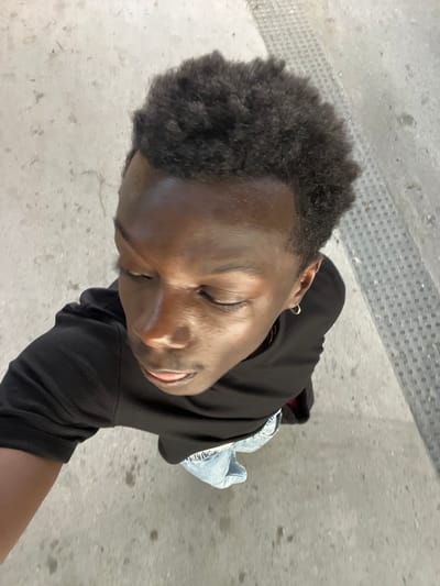 This media file features a close-up, overhead shot of a young Black person. They are wearing a black hooded sweatshirt and light-wash denim jeans. The person has short, dark, curly hair and is wearing a hoop earring in their visible ear. Their eyes are closed, and they have a slight smile on their face, suggesting a relaxed or content emotion. The lighting indicates it is daytime, and the scene appears to be outdoors on a concrete surface. In the background, there is a textured strip of gray material with a pattern of small circles, possibly a grate or textured paving. The ground surface is speckled with dirt and debris. The overall setting is urban and appears to be a public space, with the location context suggesting Sant Adrià de Besòs, Spain. No visible text is present in the image.