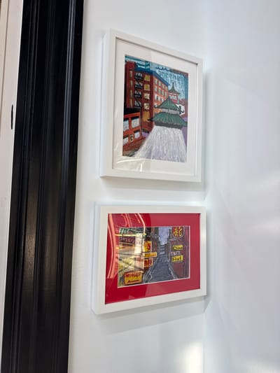 Two framed artworks are vertically mounted on a white wall. To the left, a black architectural trim is partially visible. The upper artwork, a portrait-oriented piece in a white frame with white matting, depicts a multi-story red-brick building adjacent to a green-tiered pagoda-style roof, indicative of the Chinatown Gate. The lower artwork, a landscape-oriented piece in a white frame with red matting, illustrates a narrow street scene with buildings and multiple illuminated signs featuring Asian characters and English text, including "Chinatown" and "China Pearl." Both artworks distinctly portray elements of Chinatown in Boston, Massachusetts, United States. Reflections are visible on the glass of the lower frame, suggesting ambient light. No people are present, and no actions or interactions are occurring within the visible scene.