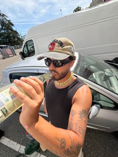 A man wearing a tan baseball cap and sunglasses is drinking from a glass bottle while standing next to a silver car in Caluire-et-Cuire, France. The man is wearing a black tank top and several beaded necklaces. He has tattoos on his right arm. In the background, a white van and trees are visible. The ground is paved with asphalt and white lines. The sky is blue with some clouds.