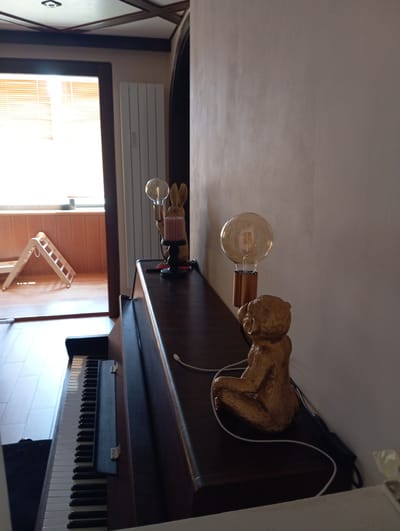 The image depicts an indoor scene, likely a domestic interior, given the presence of a piano and home decor elements. In the foreground, the edge of a dark wood piano is visible, with a section of its black and white keys exposed. Resting on the piano is a small, golden-colored statue of a monkey. Next to it, a vintage-style light bulb with a visible filament sits on a copper-colored base, which is attached to a black stand that also holds a pink candle. Further back on the piano is another decorative lamp featuring a golden rabbit figure, a bulb, and a black base. To the left of the piano, a white radiator is mounted on a wall, and beyond that, a window with slatted blinds is visible, suggesting a room with natural light. A white, wooden, A-frame child's climbing toy is set up near the window. The walls appear to be a neutral, light color, and the ceiling has a dark wood lattice design. The overall impression is of a cozy and somewhat eclectic living space. The image does not contain any individuals or provide any direct clues about its specific location within Baden, Austria.