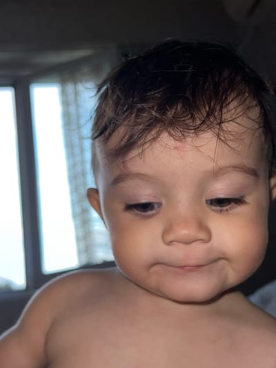 The media file captures a close-up, candid portrait of a baby, likely an infant or young toddler, in an indoor setting during the daytime in Elena, Bulgaria. The baby is shirtless, with fair skin, brown eyes, and dark, slightly messy hair that appears somewhat damp, perhaps from a bath or general activity. A small, faint red mark is visible on the baby's forehead above the left eye.

The baby's head is tilted at an angle, giving the image a dynamic feel. The baby looks towards the viewer with a curious and slightly playful expression, featuring a subtle smile or smirk. The background is out of focus, revealing a blurred bright light source, which appears to be a window, suggesting natural light entering the room. This light source creates a contrast, illuminating the baby's face, particularly the left side. The blurry horizontal lines in the background hint at blinds or window panes. The overall mood of the image is joyful and innocent. There is no visible text in the image.