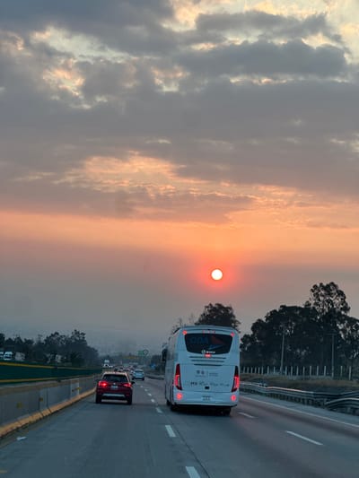 A photograph captures a sunset over a highway, featuring a bright sun in the upper-right. Heavy cloud cover stretches across the left, contrasted by the sun's orange glow. A white bus occupies the right lane, with other vehicles visible further ahead. The highway's infrastructure, including guardrails and road markings, are also visible. Trees line the edges of the highway.