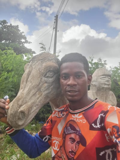 A young Black man is taking a selfie with a concrete sculpture of a horse's head in the foreground. He is wearing an orange and blue long-sleeved shirt with a repeating pattern of the Chanel logo and a portrait of Tupac Shakur. The sculpture is textured and weathered, with visible cracks. In the background, out of focus, is another concrete sculpture of a human head with closed eyes. The scene appears to be outdoors, with lush green foliage and a utility pole with wires visible in the upper right corner. The sky is partly cloudy with patches of blue. The overall setting is consistent with an outdoor art installation or public space in Egbagu, Nigeria.