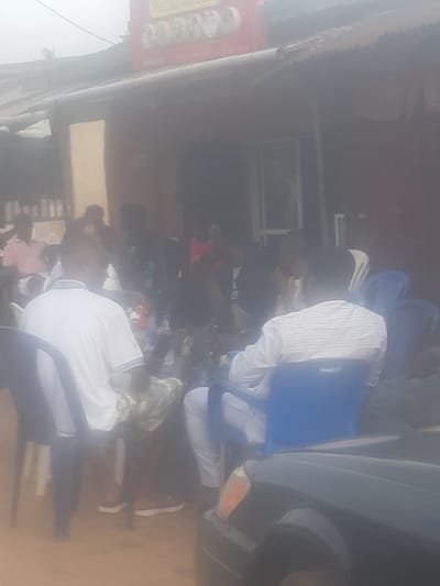 A group of people are seated around tables outside a building, possibly a restaurant, in Enugu, Nigeria. The building has a red sign with some text on it, which is difficult to read due to the blurriness of the image. Most of the people are facing away from the camera. One man in the foreground is wearing a white t-shirt with a blue collar and stripes on the sleeves. Another person is wearing a white and blue striped shirt. A dark-colored car is parked in front of the building. The overall lighting suggests it is daytime.