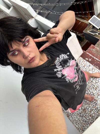 A young person with dark, choppy hair and dramatic eye makeup is taking a selfie. They are wearing a black t-shirt with a pink graphic design and bright pink shorts. The person has a nose ring and is making a peace sign with their right hand. They are standing on a surface covered in small, light-colored pebbles. In the background, there are glimpses of buildings, string lights, and two small solar panels. The lighting suggests it is daytime, likely outdoors. The image appears to be taken from a high angle, looking down at the subject. There are no clear indications of the specific location within บ้านกาญจน์กนก 23, Thailand, beyond it being an outdoor area with a paved or cobbled surface and nearby structures.