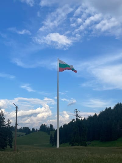 The image depicts a Bulgarian flag flying from a tall flagpole against a bright blue sky dotted with white clouds. The flag, composed of three horizontal stripes of white, green, and red, is caught by the wind, giving it a dynamic appearance. In the background, a dense forest of dark green pine trees lines a rolling green hillside. The setting appears to be a natural landscape, likely in a rural or mountainous area. A simple wooden pole with insulators, possibly part of an old electrical or communication line, is visible in the lower right portion of the image. The lighting suggests it is daytime, with the sun illuminating the scene and casting shadows. The overall mood is serene and patriotic, capturing a moment of national pride in a scenic outdoor environment. The location is identified as Smolyan, Bulgaria. There is no visible text in the image.