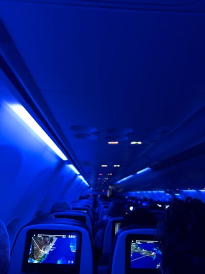 The image shows the interior of an airplane cabin bathed in blue light. A row of passenger seats is visible, each equipped with a personal entertainment screen displaying a map-like interface. The airplane wall with ventilation controls is on the left. There are no people visible in the scene besides the blurry head of a passenger in the corner. There is no information provided in the image to indicate it is in Naples, United States.