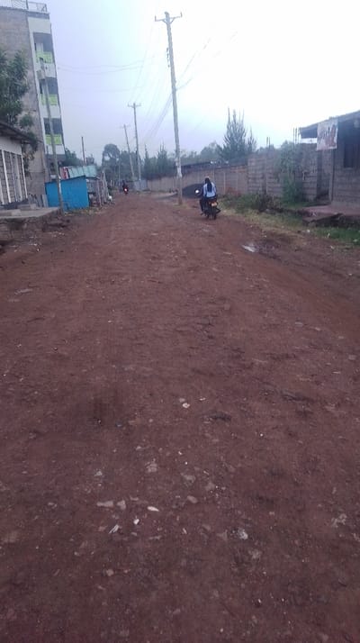 An unpaved, reddish-brown road stretches into the distance, flanked by buildings and a concrete wall. Utility poles line the street. Two motorbikes are visible, one with a rider wearing a light-colored shirt. Patches of vegetation grow alongside the road and behind the wall. The sky appears overcast, lending a muted tone to the scene. The road surface is rough and uneven.
