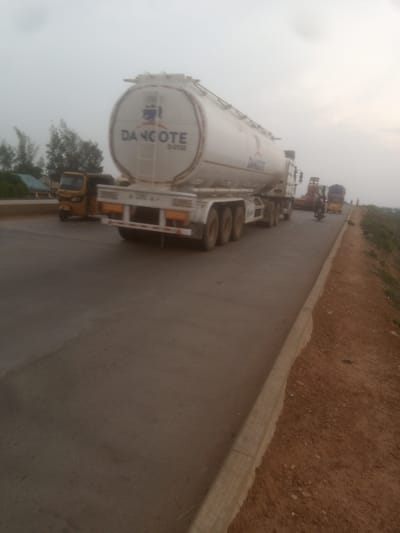 A long white tanker truck with "DANGOTE" written on its side is parked on a paved road. The truck has multiple axles and a large cylindrical tank. To the left of the tanker truck, a yellow three-wheeled vehicle is visible. In the background, there are trees and structures, suggesting an outdoor setting, possibly in a semi-urban or rural area. The sky is overcast and hazy. Further down the road, a motorcycle and another truck are present. The road itself appears to be in good condition, with a dirt shoulder on the right side. The overall lighting suggests it might be late afternoon or early morning.