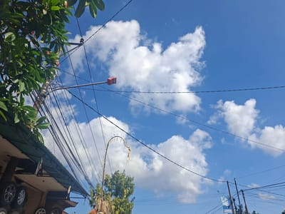 The image depicts a street scene in Blahbatuh, Indonesia, under a bright blue sky with scattered white clouds. A cluster of electrical wires dominates the foreground, strung between utility poles. To the left, a portion of a building with a green corrugated metal roof is visible, displaying stacks of car tires. A lush green tree with pink flowers overhangs the scene from the upper left. In the center of the frame, a street lamp with a pink fixture is mounted on a pole. Further to the right, a tall, curved decorative structure, possibly an offering or a temporary installation, hangs from a wire, adorned with what appears to be dried plant material and small lights. Another utility pole with more tangled wires is visible in the lower right, and a blue banner with white text is attached to it. The overall impression is of a typical urban street with overhead infrastructure and foliage.