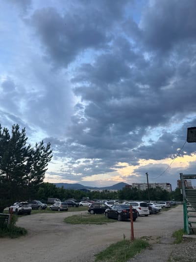 The image depicts an outdoor parking lot with several vehicles parked on a dirt and gravel surface. The parking lot is situated in front of a line of trees and buildings, with mountains visible in the background. The sky is overcast with dramatic clouds, and there is a hint of sunset light near the horizon. A large pine tree is prominent on the left side of the frame. There are no people visible in the image. The overall scene suggests a somewhat rural or suburban setting on the outskirts of a city, likely Sayanogorsk, Russia, given the typical architecture and landscape.