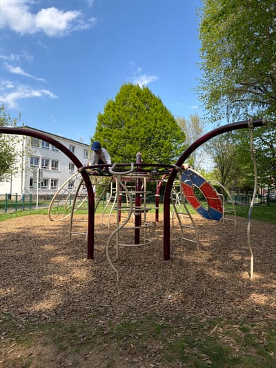 This image depicts an outdoor children's playground in Pétange, Luxembourg, under a clear, sunny sky. The central subject is a colorful metal climbing structure, featuring dark red main supports and lighter beige or white climbing bars and netting. A prominent design element is a large, round, ring-shaped piece within the structure, painted in alternating sections of red and blue.
A child, dressed in a light-colored top and a blue cap, is actively climbing on the lower section of the playground equipment. The ground beneath the play area is covered extensively with wood chips, providing a cushioned surface, while green grass borders the playground to the right.
In the background, a multi-story building with a light-colored facade and numerous windows is visible, suggesting a nearby residential or public building. Lush green trees with dense foliage surround the playground, casting shadows onto the wood chip ground, indicating bright sunshine. The sky is a vibrant blue with a few scattered white clouds and a faint contrail from an aircraft visible high above. The scene portrays a lively and well-maintained outdoor play environment. No discernible text is visible in the image.