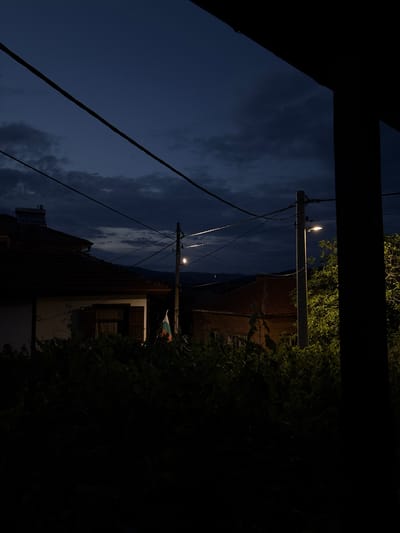 The image depicts a nighttime scene in Strelcha, Bulgaria, characterized by a twilight sky with scattered clouds. A residential building with a red-tiled roof is visible on the left, featuring a white facade and an open window with shutters. A Bulgarian flag is displayed on a flagpole. Another brick building with windows is partially visible on the right. Overhead power lines traverse the scene, supported by utility poles. A street lamp illuminates a section of the right side of the image, casting a warm glow on surrounding greenery. The foreground is dominated by dark foliage, obscuring the ground level. The overall ambiance suggests a quiet, rural evening.