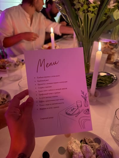 In a dimly lit indoor setting, likely a restaurant or event space, a hand holds a pink menu with "Menu" printed in elegant script at the top. The menu lists various food items in Russian, including "Хлебная корзина с сетом масел" (Bread basket with a set of oils), "Крабовый риет" (Crab rillettes), "Брускетта с печеным перцем и анчоусами" (Bruschetta with baked pepper and anchovies), "Спарка с хамоном" (Sparka with ham), "Гранд салат с кейлом и печеной свеклой" (Grand salad with kale and baked beetroot), "Трюфельный хумус с грибами" (Truffle hummus with mushrooms), "Ассорти цветных томатов" (Assortment of colored tomatoes), "Вафли с креветочным корн чизом" (Waffles with shrimp corn cheese), "Тунец с пюре из цветной капусты" (Tuna with cauliflower puree), "Морковь из печи с кремом" (Carrot from the oven with cream), and "Секретный десерт" (Secret dessert). A black and white line drawing of a table setting with cutlery and a small vase of flowers is visible on the lower right portion of the menu.
The background shows blurred figures of people dining, with a large bouquet of green flowers and white calla lilies in a vase placed on the table. Two white candles in glass holders are lit, casting a warm glow. In the foreground, a plate of food, appearing to be a salad or appetizer with creamy components and mushrooms, is partially visible, along with a fork. The overall ambiance suggests a formal or celebratory meal. The image does not provide any specific geographical indicators to place it in Chang Phueak, Thailand.