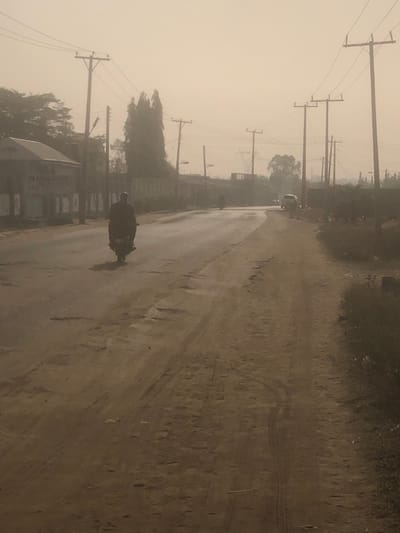 The image depicts an outdoor scene in Hanwa, Nigeria, characterized by a pervasive light brown atmospheric haze, likely dust or harmattan. The foreground shows a worn, two-lane road environment. The left lane is paved asphalt, exhibiting numerous potholes and reflective patches. The right side features an unpaved dirt path, also uneven, with visible tire tracks.
In the mid-ground of the paved road, a single individual on a dark motorcycle is observed from behind, riding away from the viewer. Further down the same road, a second, less distinct motorcycle and rider are visible, also moving away. On the far right side of the road, in the background, a light-colored vehicle (possibly an SUV) is parked or stationary, facing the direction of the camera.
Along the left side of the paved road, a single-story building with a light-colored roof is present, featuring indistinct lettering on its facade. Numerous wooden utility poles with power lines line both sides of the road, receding into the hazy distance. Behind the structures on the left, a continuous wall is visible, along with several trees, including a cluster of tall, slender trees. The right side of the dirt path is bordered by sparse, low-lying vegetation and scattered debris. The overall environment suggests an urban or semi-urban street under hazy conditions.