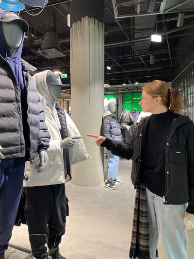 A woman points downward at a mannequin wearing a white puffy jacket. Above the mannequin is another mannequin with a jacket, jeans, and black and white sneakers. Behind the woman is a plaid scarf. The scene is in a store with a tall, cylindrical pillar with black paint at the top. The store's ceiling shows lighting fixtures and other equipment.