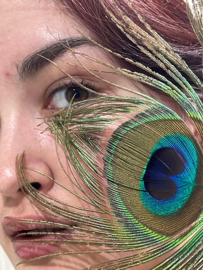 A close-up, vertical photographic portrait depicts the right side of an individual's face, located in Neftekamsk, Russia. The subject has fair skin, with a minor blemish observed superior to the right eyebrow. The right eye is open, displaying a dark brown iris and a visible pupil, directed slightly supero-laterally. Dark, defined eyelashes are present. The lips are slightly parted. Reddish-brown hair is visible along the superior-right frame edge. A peacock feather is positioned over the face. Its prominent iridescent ocellus, featuring concentric rings of blue, green, and gold, is located inferior and lateral to the right eye, partially covering the cheek. The feather's filamentous barbs extend across the right eye, the nasal bridge, and obscure portions of the upper and lower lips. The immediate background is a uniformly light, out-of-focus surface.