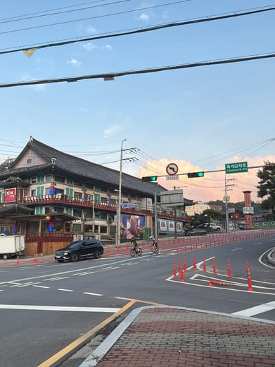 This image captures a street scene in Pocheon-si, South Korea, under a sky filled with scattered clouds and a soft, pinkish hue from what appears to be sunset. In the foreground, a paved road with clear lane markings and a pedestrian walkway made of brick pavers is visible. A black SUV is turning at an intersection, while two cyclists are riding side-by-side in the lane.
Dominating the background is a traditional Korean-style building with a dark tiled roof, multiple stories, and balconies adorned with red railings and green shutters. Signs in Korean are displayed on the building's facade, one of which features an image of meat. Adjacent to the building, traffic lights are illuminated green, indicating traffic flow. A street sign indicates "Chukseok Jct" in English, confirming the location. Construction barriers and traffic cones are placed along the roadside, suggesting ongoing road work or traffic management. The overall environment appears to be a bustling urban area with a blend of traditional architecture and modern infrastructure.