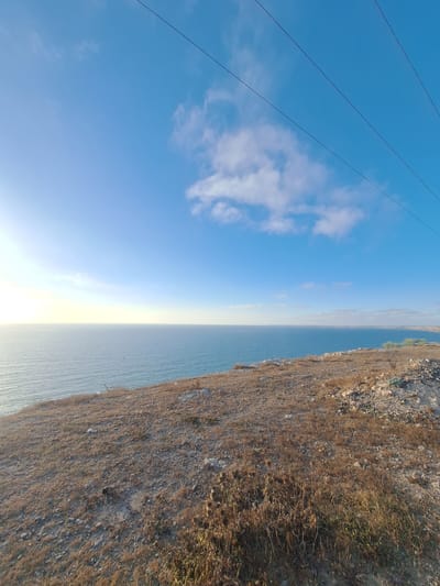 This media file displays a panoramic view of a coastal landscape. The scene is divided vertically, with the left side dominated by a bright blue sky and a few scattered white clouds. Three dark power lines are visible diagonally across the sky, suggesting human infrastructure.

The right side of the image features a rugged, dry cliff face covered in sparse, brown vegetation and scattered rocks. Below the cliff, a calm, deep blue body of water stretches to the horizon, meeting a hazy, bright sky that suggests either sunrise or sunset. The overall impression is one of a remote, natural environment.

The time of day is likely morning or late afternoon due to the low angle of the sun indicated by the bright haze on the horizon. The weather appears clear and dry, consistent with a Mediterranean or arid coastal climate. The location cues, such as the type of vegetation and the rugged coastline, are characteristic of regions like Ahrara, Morocco. No people or specific objects are identifiable, and no text is visible in the image. The overall mood conveyed is one of serenity and natural beauty.