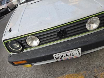 The image displays the front-end of a white Volkswagen Golf Mk2, identified by its grille design and headlight configuration. The vehicle is located in Naucalpan de Juárez, Estado de México, evidenced by the "EDOMEX" designation on its license plate. The car's white hood is visibly speckled with dust and features an aftermarket chrome hood pin on the driver's side. The black horizontal slat grille incorporates a black Volkswagen emblem at its center and is accented by thin, bright green horizontal stripes along its top and bottom edges. The passenger-side headlight is a standard round clear-lens unit, while the driver's side features a projector-style lamp set within a black ring, indicating a potential modification. Below the front bumper on the driver's side, an orange rectangular turn signal and a lower yellow fog light are visible. The textured black plastic bumper, also dusty, prominently displays a Mexican license plate with the alphanumeric identifier "MHM-516-A." The vehicle is situated on gray asphalt pavement, which exhibits a yellow painted line in the lower right quadrant of the frame. A blurry, tan-colored sedan is partially visible in the background to the upper left. No individuals or active interactions are present in the scene.