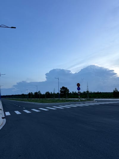This is a photograph of an asphalt road with white markings, running alongside a grassy area and a line of trees. A large, dramatic cloud formation dominates the sky, suggesting active weather. Several streetlights are visible along the road, indicating it's a developed area. Two traffic signs are mounted on a pole: a circular sign with a red border and a horizontal red bar, and a circular blue sign with a red border and a diagonal red bar. The image appears to be taken during the daytime, given the brightness of the sky. The rotation of the image suggests it was taken vertically and then turned sideways. Based on the provided location context, this scene is likely in Mui Ne, Vietnam. There is no visible text in the image.