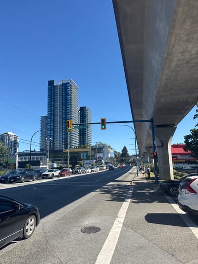 This is a ground-level, eye-level, medium shot of a sunny day on a multi-lane road in Burnaby, Canada, likely near the Metrotown area. The viewpoint is from the right lane of the road, looking north. The road is in good condition, with clear lane markings and a white crosswalk in the foreground. Several cars are visible, including a black sedan on the left, a white SUV and a red sedan further up, and a line of cars in the right lane. A pedestrian walkway runs alongside the road, with a few people walking on it. A large elevated concrete structure, part of an overpass or SkyTrain line, dominates the right side of the frame, casting a shadow onto the road. Traffic lights are visible, with one displaying a green light and another showing a red hand symbol. In the background, tall residential buildings and commercial establishments line the street. The sky is a clear, bright blue. The overall impression is of a busy urban environment on a pleasant day.