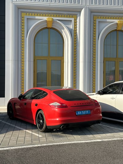 A red Porsche Panamera, seen from the rear three-quarter angle, is parked in a paved lot in Kapitan Andreevo, Bulgaria. The vehicle features a black roof, black multi-spoke wheels, and dual exhaust pipes. Its rear license plate, clearly visible, displays "XH 5019" in blue lettering on a white background, with "BG" in the top left corner and Cyrillic text "AA KAPAМАНСКИ" below the number. The car is positioned within a white-lined parking space on grey rectangular interlocking pavers. The right side of the car is partially obscured by shadows, while its left side is illuminated by direct sunlight.
To the right of the red Porsche, a white vehicle, likely an SUV or sedan, is partially visible, showing its front passenger side and a black side mirror.
In the background, a stately building with a white facade rises prominently. The building's architecture is adorned with gold accents and intricate details. It features multiple vertical pilasters with subtle fluting and gold rectangular insets running along their length. The upper part of the facade includes a frieze with a repeating geometric pattern. Two large arched doorways or windows, framed by thick white architraves, are central to the building's design. Each arch is further outlined by a golden frame with a stepped, tooth-like pattern, and capped with a distinctive golden, trapezoidal decorative element at its apex. The doorways themselves have golden frames dividing them into multiple panes. The building's white surfaces are brightly lit by direct sunlight, creating strong contrasts with the golden accents and casting sharp shadows. The immediate foreground shows a strip of dark grey asphalt. No people are visible in the scene.
