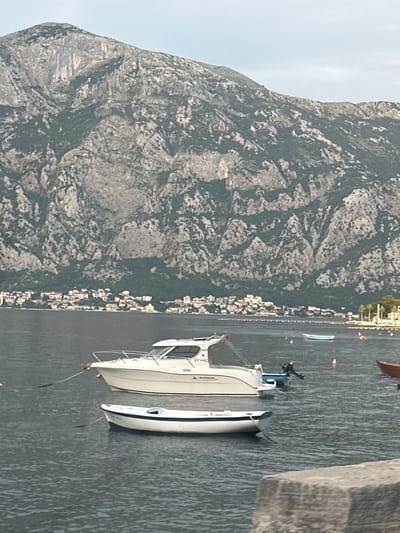 In Prčanj, Montenegro, on a body of water, two boats are moored. The larger boat, a white cabin cruiser with a navy blue stripe, is in the foreground, tethered to the left. A smaller, white rowboat with a navy stripe is positioned below and slightly to the right of the cabin cruiser, also moored. In the background, the town of Prčanj stretches along the coastline, with buildings featuring red-tiled roofs nestled at the base of a rugged, green-covered mountain. The water is calm, and the sky above is a pale blue with some cloud cover.