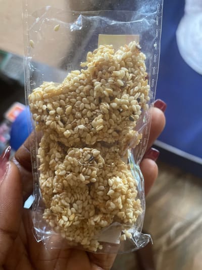 A close-up shot focuses on a person's hands holding a clear plastic package containing small, light-colored sesame seed candies. The candies appear to be slightly irregular in shape and are clustered together within the package. Some dark specks, possibly dirt or stray sesame seeds, are visible among the lighter ones. The person's fingernails are painted a dark red color. The background is blurred, showing hints of a blue surface and other indistinct objects, suggesting an indoor setting. There is no information in the image to indicate the location is Kakau, Nigeria.