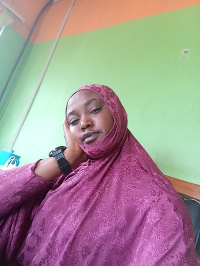 A woman wearing a burgundy hijab and a black smartwatch rests her head on her hand, posing for a selfie. The background features a two-toned wall, with the top section painted orange and the lower section painted green. A white pipe runs vertically along the wall. A turquoise container with white straps rests on a surface to the left of the woman. The image location is not apparent from the information.
