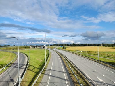 The image captures a highway interchange on a clear day with scattered clouds. The scene is set in Edise, Estonia, as indicated by the blue road sign displaying "Lihula 17 km". The highway has multiple lanes and is bordered by green grassy areas and a field of yellow crops. Several streetlights are visible along the road, and guardrails line both sides. The sky is a bright blue with dynamic cloud formations, suggesting it is daytime. No people or vehicles are present in the frame, giving the impression of a quiet, undeveloped landscape. The overall environment is one of modern infrastructure integrated into a rural setting.