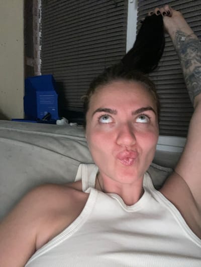 A woman, with dark hair and a tattoo on her right arm, is lying down and holding a portion of her hair above her head. She is wearing a white tank top and has her lips pursed, looking upwards. The background appears to be a room with grey blinds and a beige couch. There is also a blue box and some white objects on a surface behind her. The image is taken from a close-up perspective, focusing on the woman's face and upper body. There is no indication of the location in the city of Мосметаллоконструкция Онт, Russia.