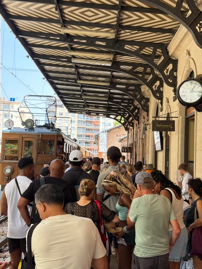 The image depicts a crowded platform at the Palma de Mallorca train station in Spain. A vintage wooden tram, identified by the number "1" on its front, is visible. Passengers are standing in line under a patterned awning supported by ornate metalwork. The station building features a cream-colored facade with arches and a large, antique clock. Signage for "Informacio" (Information) is also present. The sky is clear and blue. The people are a diverse group, with many wearing casual summer clothing. Some individuals are looking towards the tram, while others are engaged in conversation. The scene conveys a sense of waiting and anticipation, likely for the tram's departure.