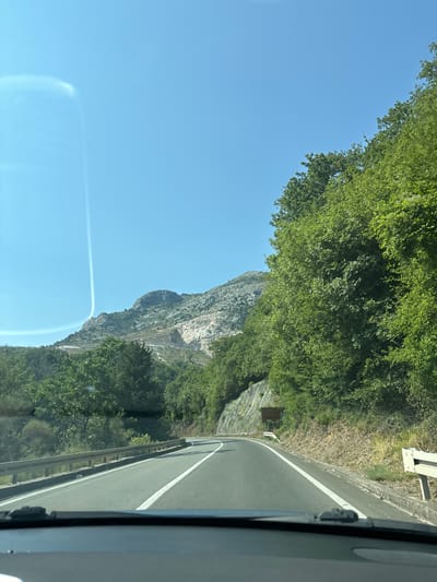 This media file depicts a view from inside a vehicle traveling along a road in Mišići, Montenegro. The scene is dominated by a clear blue sky and lush green foliage. To the left, a sheer, rocky mountainside rises dramatically, with patches of greenery clinging to its steep slopes. The road itself is asphalt with white lane markings and a metal guardrail along the side.
The time of day appears to be daytime, likely late morning or early afternoon, given the bright sunlight casting shadows. The weather seems fair and sunny. A sign is visible on the right side of the road, partially obscured by trees, but appears to be a directional or informational sign. The perspective suggests a scenic drive through a mountainous, forested region. The overall impression is one of a journey through a natural landscape.