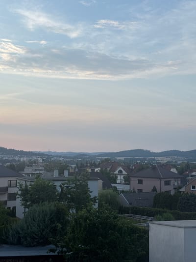 The image captures a panoramic view of a European town, likely Strakonice, Czechia, from an elevated perspective. The scene is divided into two distinct halves by the horizon. The left side is dominated by a vast sky filled with wispy, cirrus clouds against a pale blue backdrop, suggesting a late afternoon or early evening time of day, possibly around sunset given the soft, diffused light. The right side of the image showcases a dense residential area nestled on a hillside. Numerous houses with red-tiled roofs are visible, interspersed with lush green trees and hedges. The architecture appears typical of Central European towns. In the distance, a rolling, tree-covered hill forms the backdrop, hinting at a natural landscape surrounding the urban area. The overall atmosphere is serene and peaceful, with no discernible activity or people. The lighting conditions and the subtle hues in the sky contribute to a tranquil mood. There is no visible text in the image.