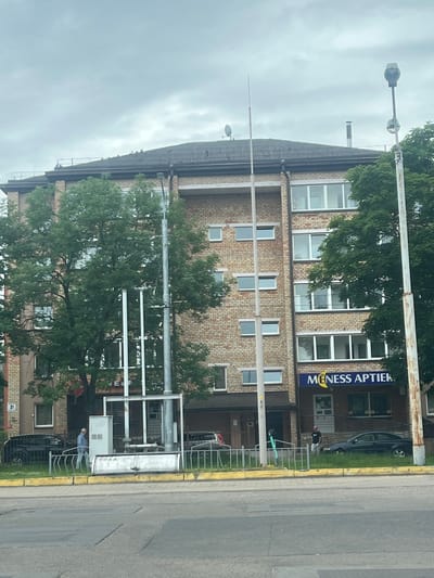 This image shows a street scene in Daugavpils, Latvia. A brick building with multiple windows lines the street, and a blue banner with white text that reads "MENESS APTIEKA" is visible on the building's facade. Several trees with lush green foliage are present, partially obscuring the building. A street light stands tall on the left side of the image, and a metal fence runs along the sidewalk. A person wearing a light blue shirt and white pants is standing near the fence, and a dark-colored car is parked in front of the building. The weather appears to be overcast, suggesting a cloudy day. The overall scene depicts an urban environment with a mix of commercial and residential elements.