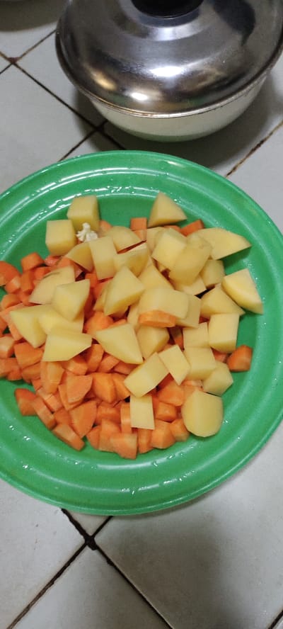 A green plastic plate filled with chopped potatoes and carrots sits on a tiled floor. The tiles are off-white and have dark grout lines. There is a silver pot lid out of focus in the background. The location is Kecamatan Lawang, Indonesia, based on the provided coordinates. The image suggests food preparation in a domestic kitchen setting. The potatoes and carrots are likely being prepared for a meal. The plate is the only subject of the image and is centered and in focus. The background is out of focus and shows only the floor. There is no sign of any people or other subjects in the image. The scene is static and depicts the food preparation process.