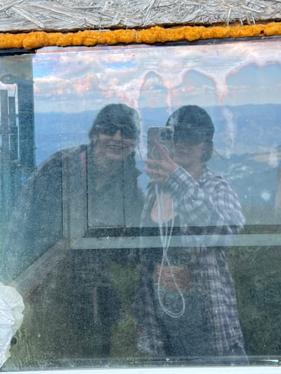 In Chorna Tysa, Ukraine, two individuals are reflected in a dirty window. On the left, a person wearing sunglasses and a dark jacket is visible. To the right, another person holds up a smartphone to take a selfie, wearing a plaid shirt and a dark baseball cap. The reflection shows a cloudy sky and a distant landscape. The window frame is visible, with orange spray foam along the top edge and some debris at the bottom left.