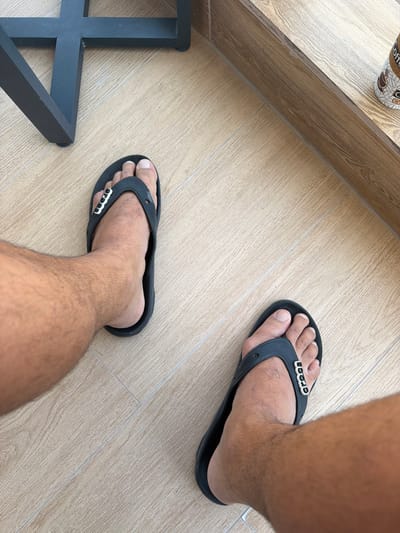 The image captures a high-angle view looking down at a person's feet and lower legs on a light-colored, wood-grain tiled floor. The person has tanned skin and visible hair on their legs, and is wearing black Crocs brand flip-flops. The white "CROCS" logo is clearly displayed on the straps of both sandals.
In the upper left portion of the frame, a disposable coffee cup with a distinctive brown and white "coffee" pattern is partially visible, resting on a raised ledge or counter that matches the wood-grain tile aesthetic. To the lower left, a dark-colored, possibly black or dark grey, metal furniture leg with a geometric, grid-like structure is partially visible on the floor.
The scene is brightly and evenly lit, suggesting it is daytime. The setting appears to be a casual indoor or covered outdoor space, such as a cafe, lounge area, or a modern home. The clean, wood-look tiled floor and contemporary furniture suggest a well-maintained environment. The given location context for this scene is Lusail, Qatar. The activity depicted is simply the person standing or walking. No specific emotions are discernible.