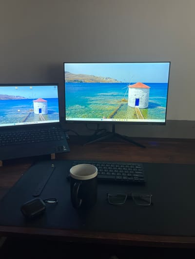The image shows a workstation setup. There are two computer screens displaying the same background of a white windmill with an orange roof overlooking the sea. A laptop is positioned on the desk, along with a separate keyboard, a pair of glasses, a black mug, and a pair of black ear buds. The scene appears to be indoors and on a dark wooden desk. There is no clear evidence connecting this workstation to Avellaneda, Argentina.