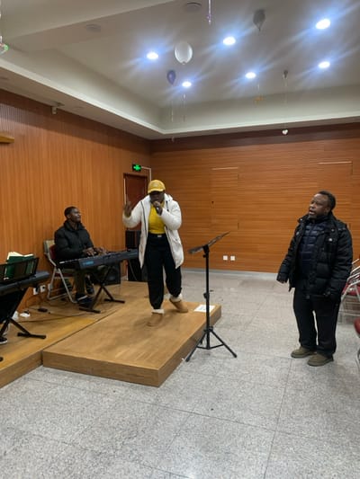 The scene depicts an indoor musical performance or rehearsal within an event room located in Shenyang City, China. The room features horizontal light-brown wood paneling on the walls, a light grey speckled tiled floor, and a white ceiling with multiple recessed circular lights. Several white and purple balloons are suspended from the ceiling.

Three individuals are present. In the center, a person, appearing to be a Black female, stands on a raised two-tiered wooden stage. She is wearing a yellow baseball cap, a yellow long-sleeved top, black pants with white cuffs, and light brown boots. She holds a black microphone in her right hand and has her left hand raised with fingers splayed, actively performing. To her immediate right, a black microphone stand is positioned on the stage.

To the left of the central performer, seated on a white chair on the lower tier of the wooden stage, is a second person, appearing to be a Black male. He is dressed in a dark puffer jacket and dark pants, and is operating a blue Yamaha electronic keyboard. A black music stand holding green folders is visible to his left.

To the right of the stage, standing on the tiled floor, is a third person, appearing to be a Black male. He is wearing a dark puffer jacket, dark pants, and olive-green shoes. He is looking towards the central performer, with his mouth slightly ajar, seemingly observing or singing along. An emergency exit sign with green text is visible on the left wall.