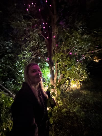 A blonde woman stands to the left of a tree trunk in a dimly lit outdoor setting, with pink lights scattered in the tree branches and yellow lights illuminating the ground to the right. The woman is wearing a dark jacket and a necklace, and her face is turned to the side. The background consists of trees and foliage. The location is Zavod Mikhailovskii, Russia.