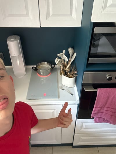 This media file is a vertical, first-person perspective image taken in a kitchen. The subject is a person, partially visible from the chest up, wearing a red t-shirt. Their head is tilted back, and their tongue is sticking out, suggesting a playful or humorous pose.

The setting appears to be a modern kitchen with white cabinets and a dark blue backsplash. Above the backsplash, there is a microwave oven and a pink towel hanging. On the counter, a white ceramic holder is filled with various kitchen utensils, including spoons and forks. A pot is placed on a white induction cooktop, which is illuminated with a red glow, indicating it is in use. To the left of the cooktop, a white cylindrical object, possibly a soap dispenser or air freshener, is mounted on the wall.

The lighting suggests it is daytime, but there are no clear indications of the time of day beyond that. The overall atmosphere is casual and lighthearted, primarily conveyed through the subject's facial expression. The location context provided is Madara, Bulgaria. No other text is visibly legible in the image.