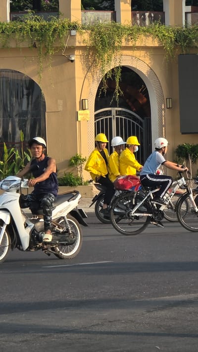 The image depicts a street scene in Unknown, Vietnam. A beige building with archways and greenery is visible on the left. On the road, a man wearing a helmet, dark t-shirt, and patterned pants is on a white motorcycle. Behind the motorcycle, there is a group of people on bicycles. Three people wearing yellow jackets and helmets are positioned directly behind the motorcycle, with another person on a bicycle further back, wearing a light blue shirt, dark pants with white stripes, and a helmet. The road is grey, suggesting it is paved.