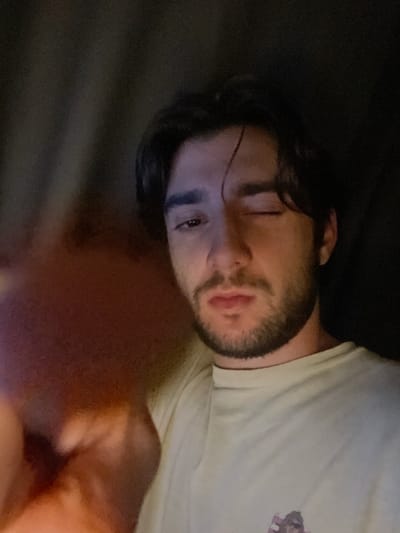 A young man with dark hair and a beard is shown in a close-up shot. He is wearing a light-colored t-shirt. His left eye is closed, and his right eye is open, looking towards the camera. The lighting is dim, with a reddish-orange glow in the foreground on the left side of the image, obscuring a portion of the man's face. The background is dark and appears to be a wall or fabric. The image was captured in Huixquilucan de Degollado, Mexico.