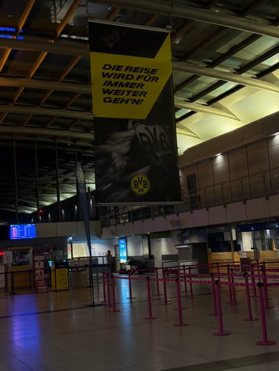 The image depicts the interior of a building, likely an airport or train station, based on the presence of check-in counters, queue barriers, and a departure/arrival screen in the background. A large yellow banner with black lettering hangs prominently from the ceiling, displaying the German phrase "DIE REISE WIRD FÜR IMMER WEITER GEH'N!" which translates to "The journey will always go on!". The banner also features the logo of Borussia Dortmund (BVB), a football club, indicated by the "BVB 09" emblem.

In the mid-ground, there are several check-in stations or service counters, some with signage. Pink queue barriers are set up, suggesting a waiting area. A trash receptacle labeled "WASTE" is visible. A person in a grey shirt is standing at one of the counters. In the background, a blue digital screen displays what appears to be flight or train information.

Further in the background, people are seated on the floor, some appearing to rest. The architecture includes exposed ceiling beams, ductwork, and large windows or mirrored surfaces reflecting the interior. The overall lighting is dim, with artificial lights illuminating the space. The floor is tiled.

Given the BVB logo and the German text, the location is very likely in Dortmund, Germany, and the banner is associated with the Borussia Dortmund football club, suggesting a transportation hub serving travelers who are fans of the club or participating in events related to it.